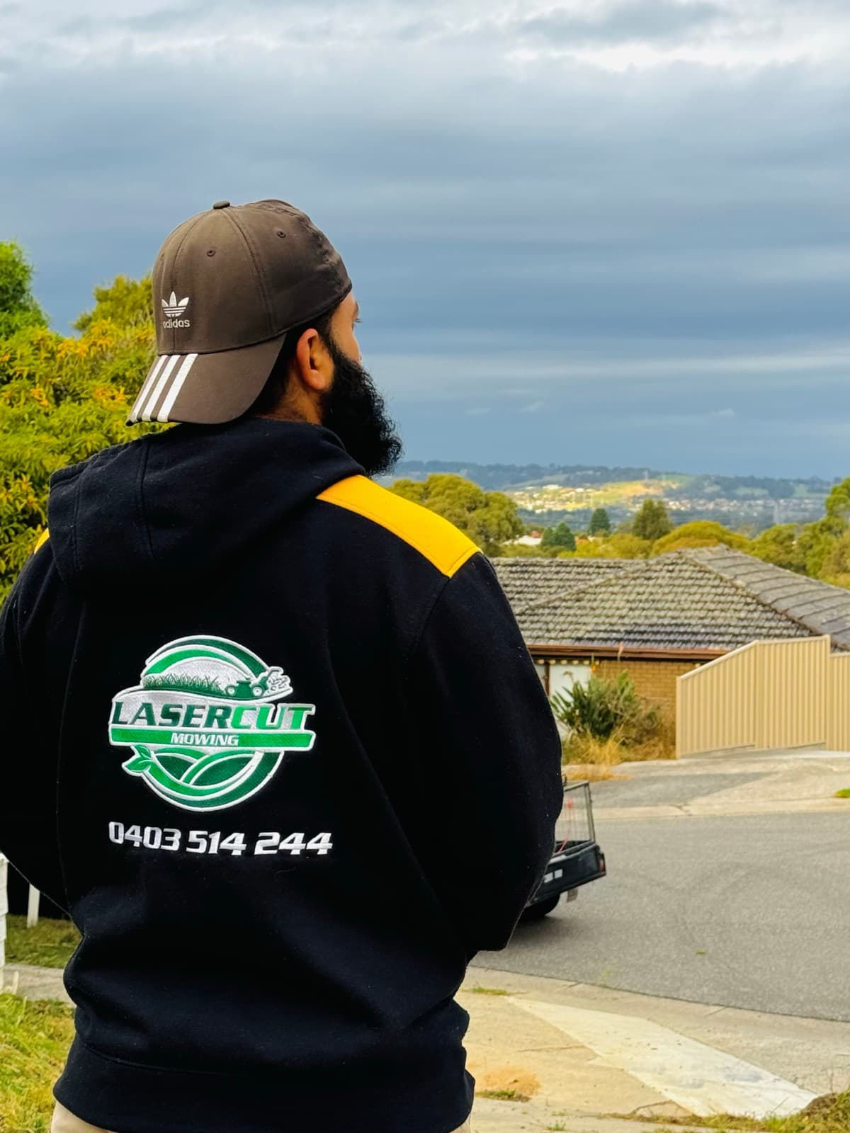 The owner of Laser Cut Mowing standing in a Cranbourne street, wearing the branded hoodie with the company logo and phone number, looking out across the suburb.